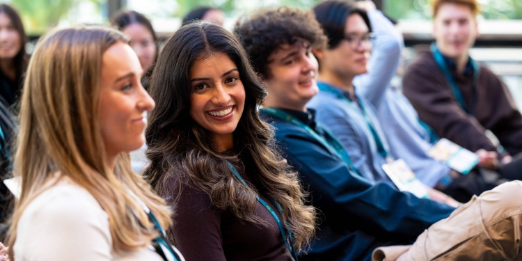 graduate woman in brown with long hair looking and smiling toward camera with other graduates sitting beside her and facing toward the front