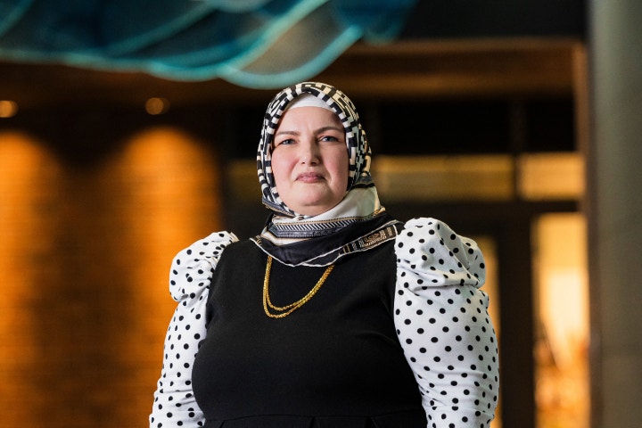 Woman wearing black dress with white dots and headscarf standing in front of ambient laneway.