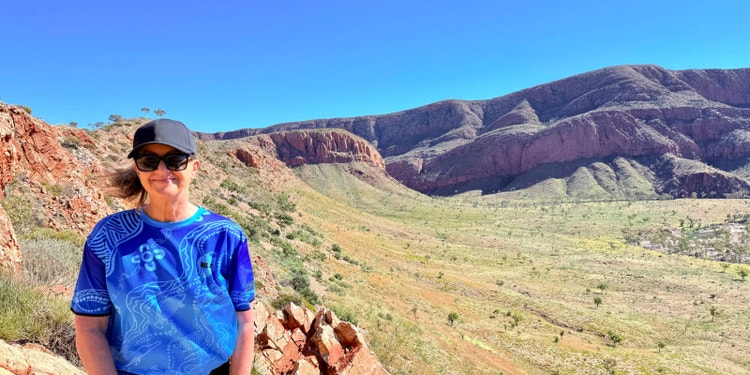 woman in blue shirt with Indigenous pattern standing in outback desert with red rocky mountain in background