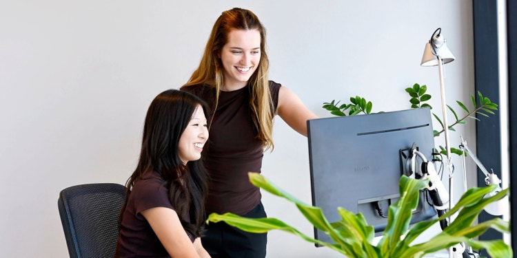 woman sitting and looking at computer with another woman standing behind her and pointing at screen while smiling