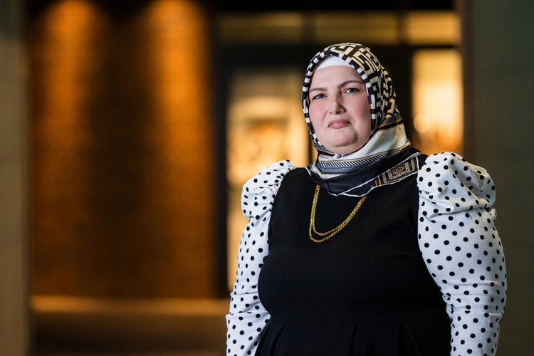 Woman wearing black dress with white dots and headscarf standing in front of ambient laneway.