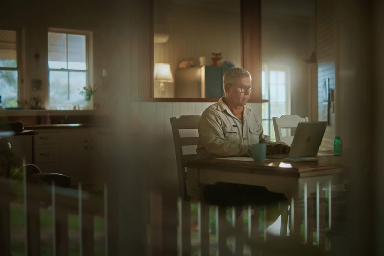 Man sitting inside at dining table looking at open laptop