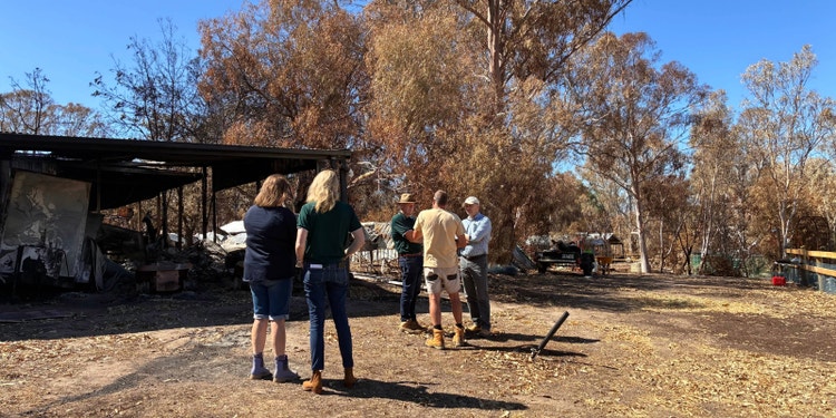 woman wearing suncorp polo standing in front of home destroyed by bushfire with customers